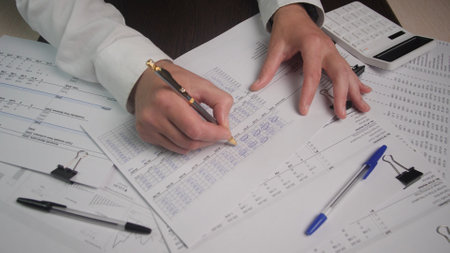 Individual is intensely reviewing and writing on financial documents, surrounded by pens and a calculator, showcasing a productive office atmosphere.の写真素材