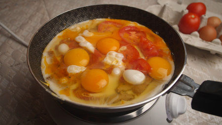 Fresh eggs are being cooked with chopped tomatoes and cheese in a frying pan on a stovetop, while fresh produce is arranged nearby, creating a warm atmosphere.の写真素材