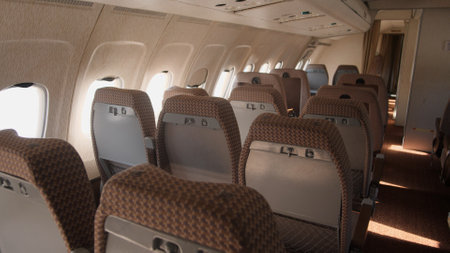 Rows of empty airplane seats fill the cabin space, illuminated by soft afternoon light coming...