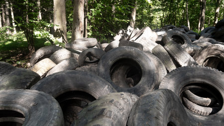 A large pile of discarded rubber tires accumulates in a natural forest setting, exacerbating...