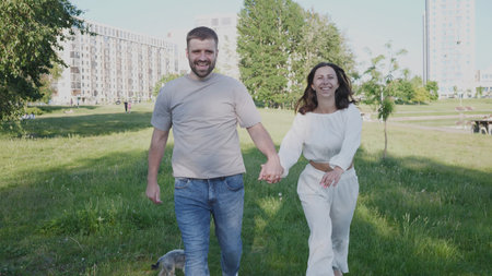 Couple Enjoying a Sunny Day While Walking Hand in Hand in a Green Park With a Dog, Surrounded by City Buildings on a Beautiful Afternoonの写真素材
