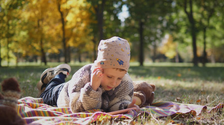 Child Enjoys Reading on a Blanket in a Sunlit Park Surrounded by Autumn Trees in Warm Colors During a Pleasant Afternoonの写真素材
