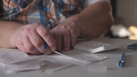 Person examines receipts and writes notes at a desk with a computer and calculator during the day in...