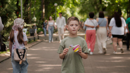 Young boy holding an ice cream cup while standing on tree-lined park path with people walking