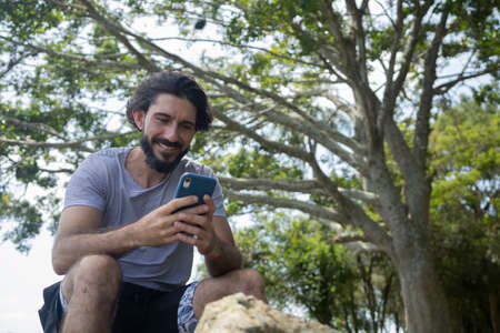 Young man at a park on a beautiful sunny day with mobile phone. Mobile phone for working and leisure. Home office concept. Green and nature background. high quality photoの写真素材