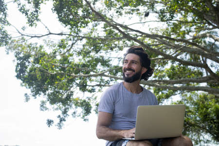 Young man at a park on a beautiful sunny day with laptop on his lap. Gray notebook for working and leisure. Home office concept. Green and nature background. high quality photoの写真素材