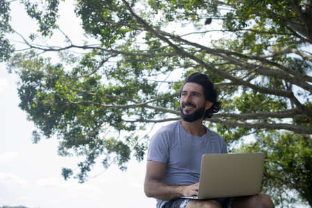 Young man at a park on a beautiful sunny day with laptop on his lap. Gray notebook for working and leisure. Home office concept. Green and nature background. high quality photoの写真素材