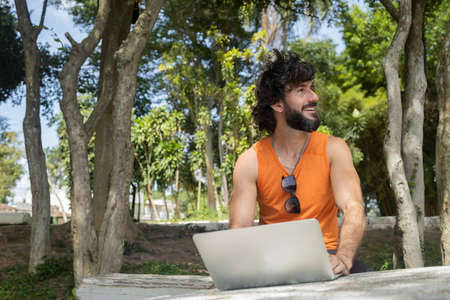 Young man at a park on a beautiful sunny day with laptop on his lap. Gray notebook for working and leisure. Home office concept. Green and nature background. high quality photoの写真素材