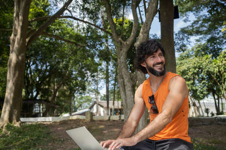 Young man at a park on a beautiful sunny day with laptop on his lap. Gray notebook for working and leisure. Home office concept. Green and nature background. high quality photoの写真素材