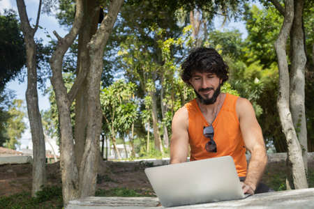Young man at a park on a beautiful sunny day with laptop on his lap. Gray notebook for working and leisure. Home office concept. Green and nature background. high quality photoの写真素材