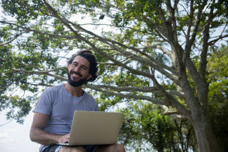 Young man at a park on a beautiful sunny day with laptop on his lap. Gray notebook for working and leisure. Home office concept. Green and nature background. high quality photoの写真素材