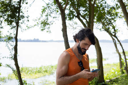 Young man at a park on a beautiful sunny day with mobile phone. Mobile phone for working and leisure. Home office concept. Green and nature background. high quality photoの写真素材