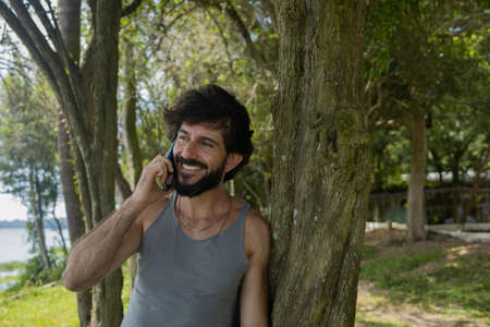 Young man at a park on a beautiful sunny day with mobile phone. Mobile phone for working and leisure. Home office concept. Green and nature background. high quality photoの写真素材