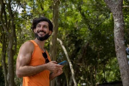 Young man at a park on a beautiful sunny day with mobile phone. Mobile phone for working and leisure. Home office concept. Green and nature background. high quality photoの写真素材