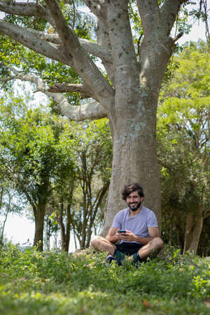 Young man at a park on a beautiful sunny day with mobile phone. Mobile phone for working and leisure. Home office concept. Green and nature background. high quality photoの写真素材