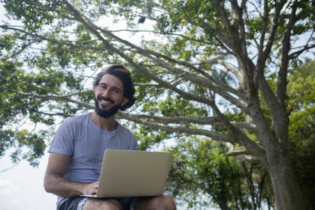 Young man at a park on a beautiful sunny day with laptop on his lap. Gray notebook for working and leisure. Home office concept. Green and nature background. high quality photoの写真素材