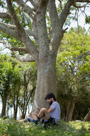 Young man at a park on a beautiful sunny day with laptop on his lap. Gray notebook for working and leisure. Home office concept. Green and nature background. high quality photoの写真素材