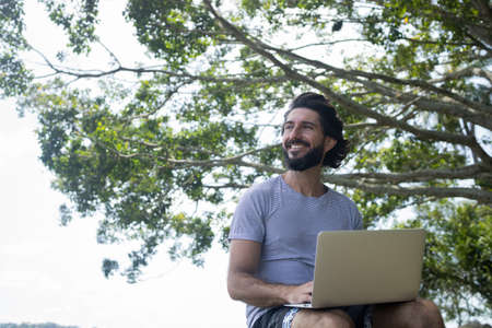 Young man at a park on a beautiful sunny day with laptop on his lap. Gray notebook for working and leisure. Home office concept. Green and nature background. high quality photoの写真素材
