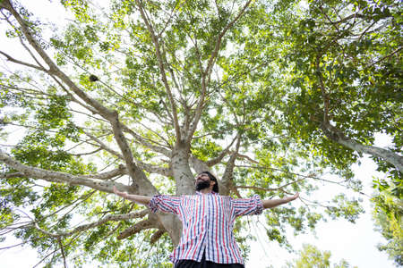 Portrait of smiling young man at a park on a beautiful sunny day. Man with open arms in front of a big a tree. Green and nature background. high quality photoの写真素材