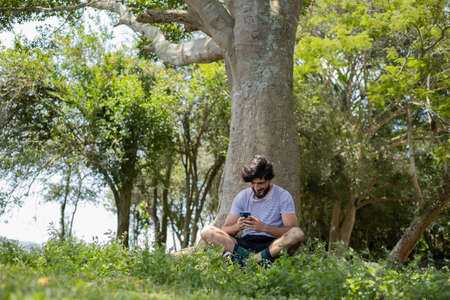 Young man at a park on a beautiful sunny day with mobile phone. Mobile phone for working and leisure. Home office concept. Green and nature background. high quality photoの写真素材
