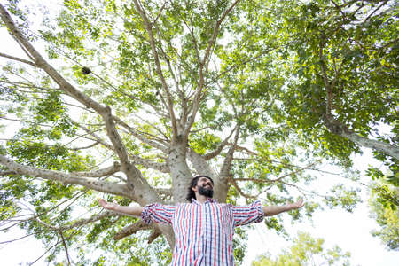 Portrait of smiling young man at a park on a beautiful sunny day. Man with open arms in front of a big a tree. Green and nature background. high quality photoの写真素材