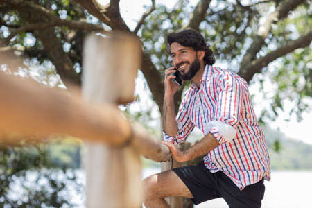 Young man at a park on a beautiful sunny day with mobile phone. Mobile phone for working and leisure. Home office concept. Green and nature background. high quality photoの写真素材