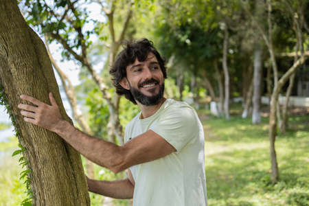 Portrait of smiling young man at a park on a beautiful sunny day. Man hugging a tree. Green and nature background. high quality photoの写真素材