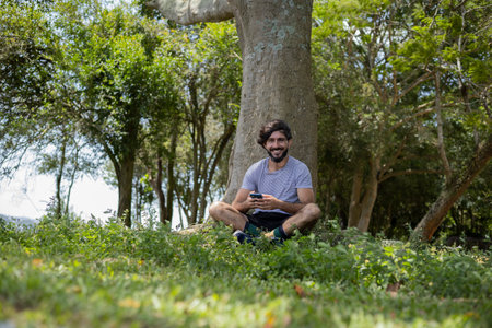 Young man at a park on a beautiful sunny day with mobile phone. Mobile phone for working and leisure. Home office concept. Green and nature background. high quality photoの写真素材