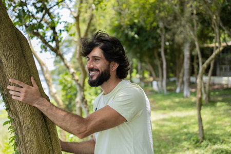 Portrait of smiling young man at a park on a beautiful sunny day. Man hugging a tree. Green and nature background. high quality photoの写真素材