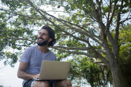Young man at a park on a beautiful sunny day with laptop on his lap. Gray notebook for working and leisure. Home office concept. Green and nature background. high quality photoの写真素材