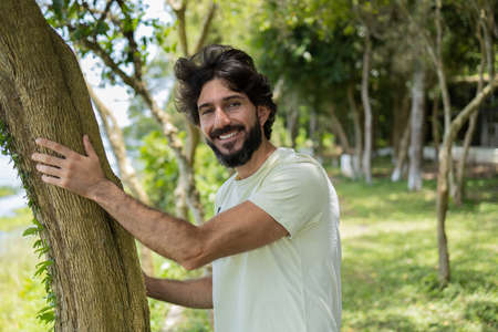 Portrait of smiling young man at a park on a beautiful sunny day. Man hugging a tree. Green and nature background. high quality photoの写真素材