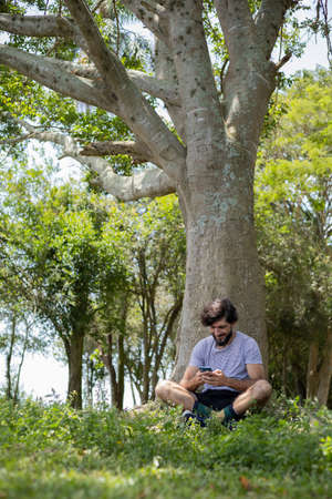 Young man at a park on a beautiful sunny day with laptop on his lap. Gray notebook for working and leisure. Home office concept. Green and nature background. high quality photoの写真素材