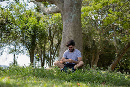 Young man at a park on a beautiful sunny day with laptop on his lap. Gray notebook for working and leisure. Home office concept. Green and nature background. high quality photoの写真素材