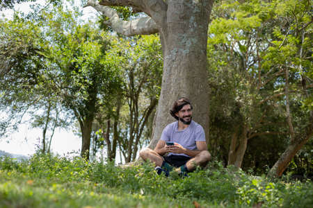 Young man at a park on a beautiful sunny day with laptop on his lap. Gray notebook for working and leisure. Home office concept. Green and nature background. high quality photoの写真素材