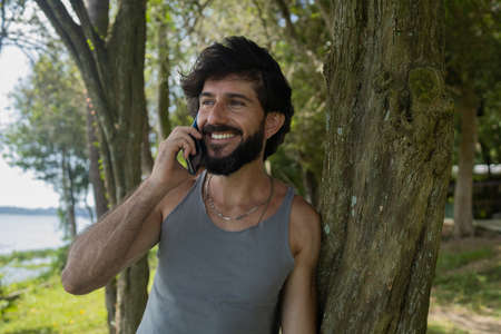 Young man at a park on a beautiful sunny day with mobile phone. Mobile phone for working and leisure. Home office concept. Green and nature background. high quality photoの写真素材