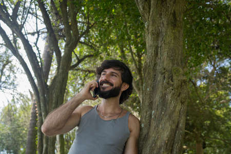 Young man at a park on a beautiful sunny day with mobile phone. Mobile phone for working and leisure. Home office concept. Green and nature background. high quality photoの写真素材