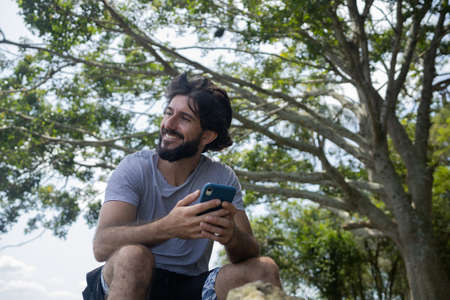 Young man at a park on a beautiful sunny day with mobile phone. Mobile phone for working and leisure. Home office concept. Green and nature background. high quality photoの写真素材
