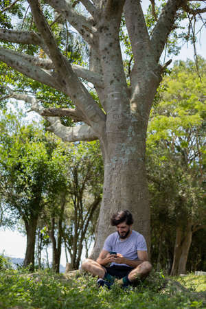 Young man at a park on a beautiful sunny day with mobile phone. Mobile phone for working and leisure. Home office concept. Green and nature background. high quality photoの写真素材