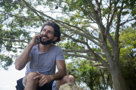 Young man at a park on a beautiful sunny day with mobile phone. Mobile phone for working and leisure. Home office concept. Green and nature background. high quality photoの写真素材