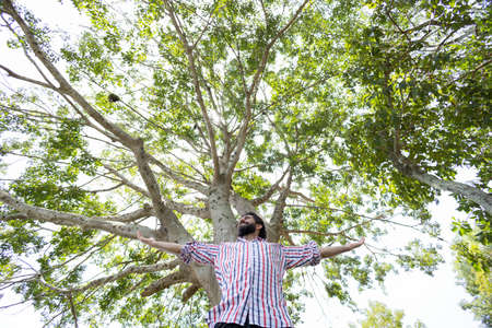 Portrait of smiling young man at a park on a beautiful sunny day. Man with open arms in front of a big a tree. Green and nature background. high quality photoの写真素材