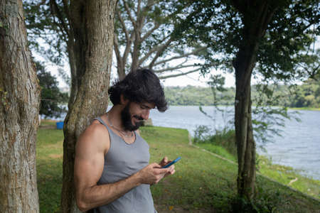 Young man at a park on a beautiful sunny day with mobile phone. Mobile phone for working and leisure. Home office concept. Green and nature background. high quality photoの写真素材