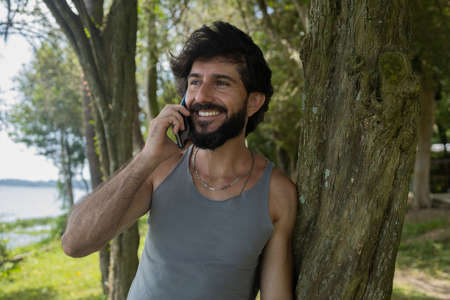 Young man at a park on a beautiful sunny day with mobile phone. Mobile phone for working and leisure. Home office concept. Green and nature background. high quality photoの写真素材