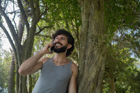 Young man at a park on a beautiful sunny day with mobile phone. Mobile phone for working and leisure. Home office concept. Green and nature background. high quality photoの写真素材
