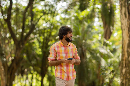 View of young man using a smartphone at day time with a green park in the background. Mobile phone,...