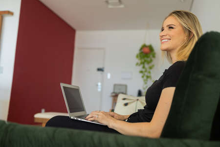 Young woman working at home from her living room couch with laptop on her lap. Home office concept. Gray notebook for working. Home office concept. high quality photoの写真素材