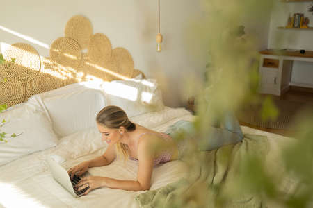 Young woman working at home from her bedroom with laptop on her bed. Woman lying down. Home office concept. Gray notebook for working. Cozy and comfortable. Home office concept. high quality photoの写真素材