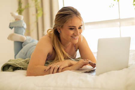 Young woman working at home from her bedroom with laptop on her bed. Woman lying down. Home office concept. Gray notebook for working. Cozy and comfortable. Home office concept. high quality photoの写真素材