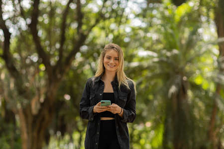 Young woman using a smartphone at day time with a green park in the background. High quality photo. Mobile phone, technology, urban concept. high quality photoの写真素材
