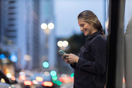 Young woman using a smartphone at night time with city view landscape in the background. High quality photo. photo. Mobile phone, technology, urban concept.の写真素材
