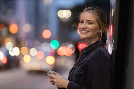 Young woman using a smartphone at night time with city view landscape in the background. High quality photo. photo. Mobile phone, technology, urban concept.の写真素材